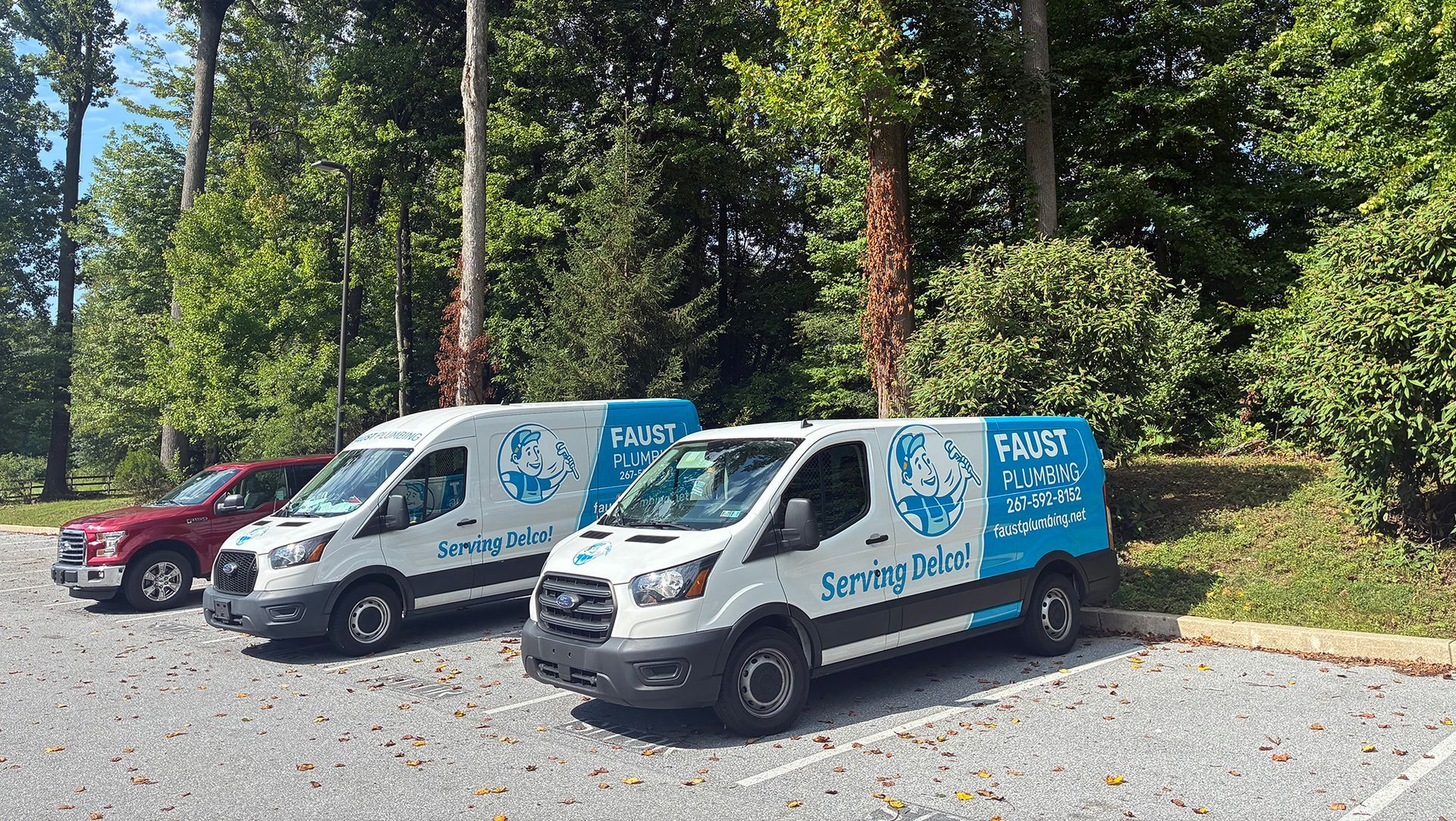 Two white service vans and a red car parked on gravel by trees, one van with blue branding.