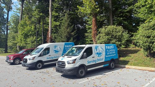 Two white delivery vans and a red car parked on a gravel lot beside trees and greenery.