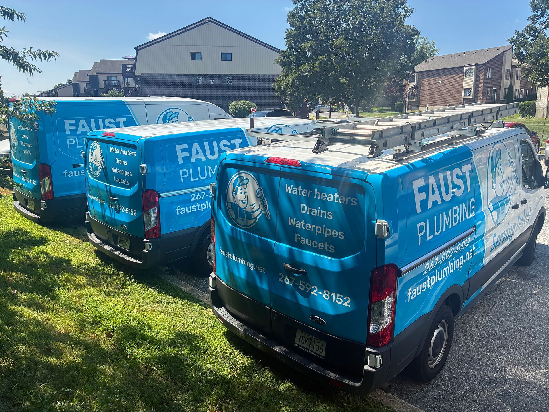 Three blue Faust Plumbing vans parked on a grassy driveway beside a house on a sunny day.