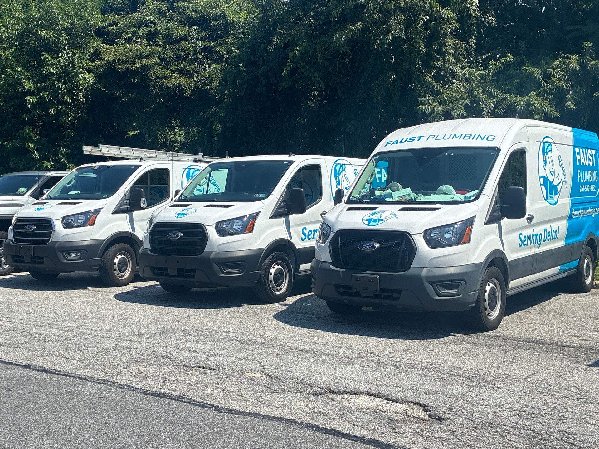 Row of white delivery vans parked on a sunny street beside trees.