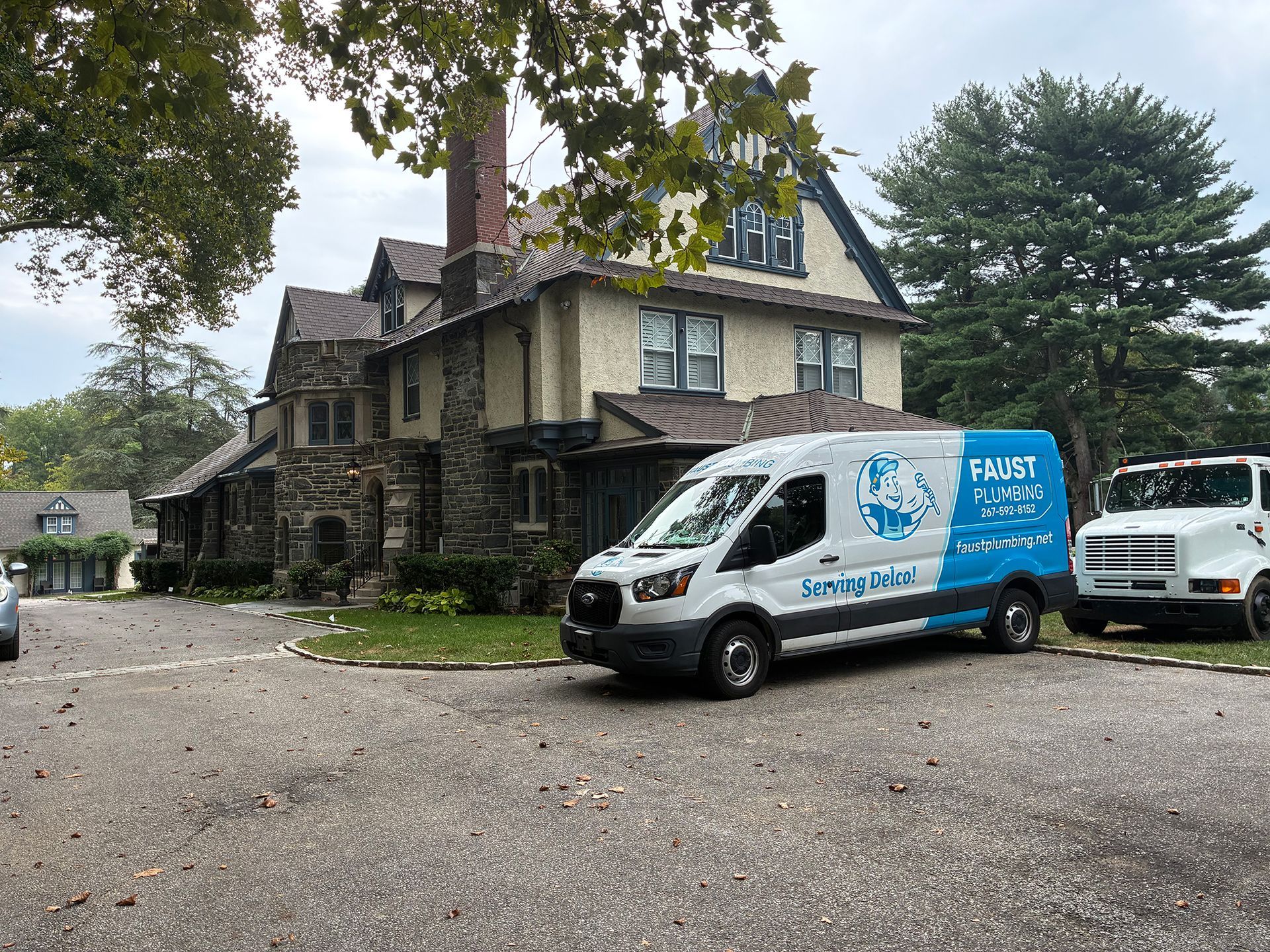 Two service vans parked beside a weathered house under trees on a gravel driveway.