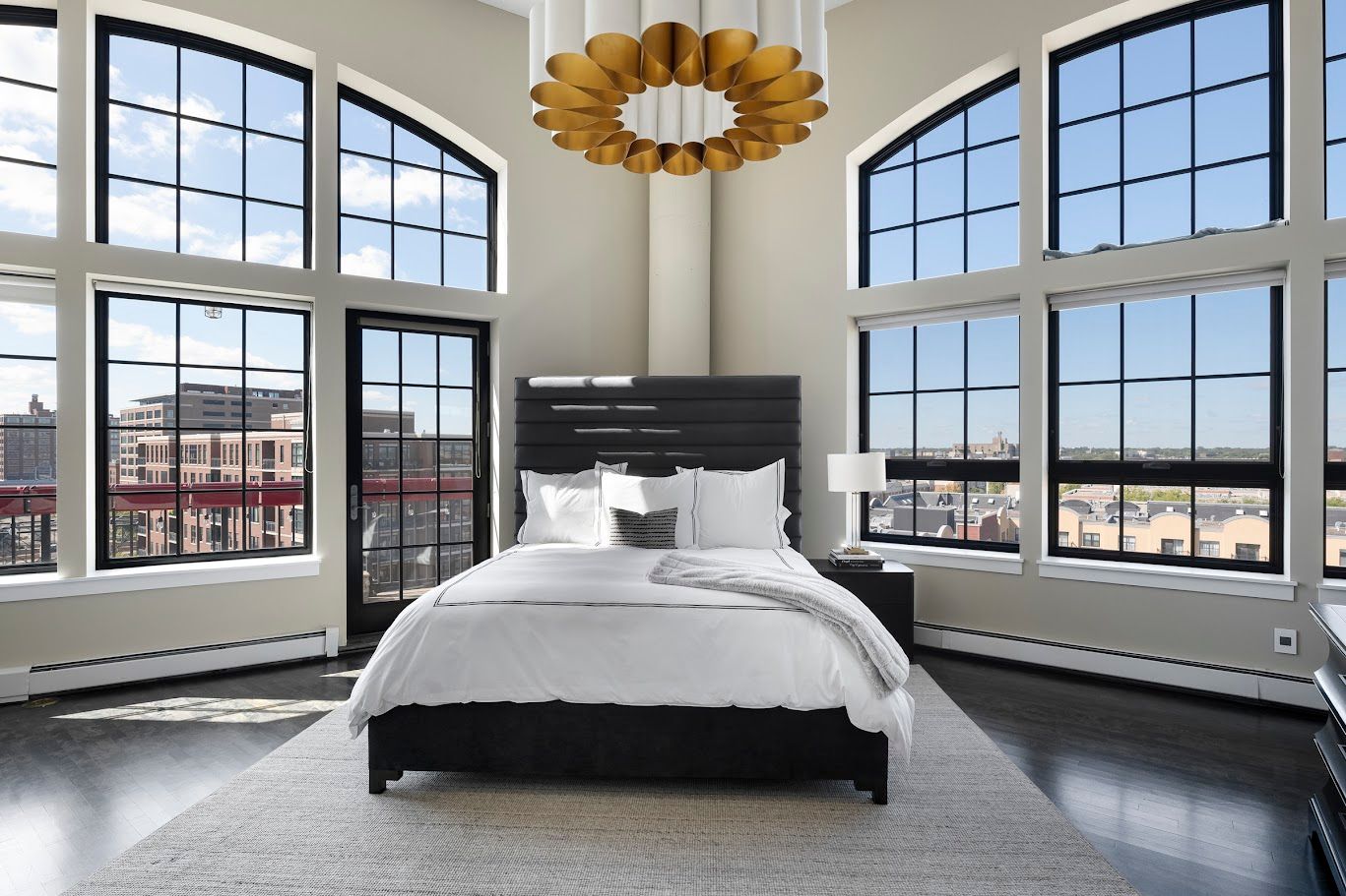Bedroom with large windows, city view, white bedding, black headboard and furniture, gold chandelier.