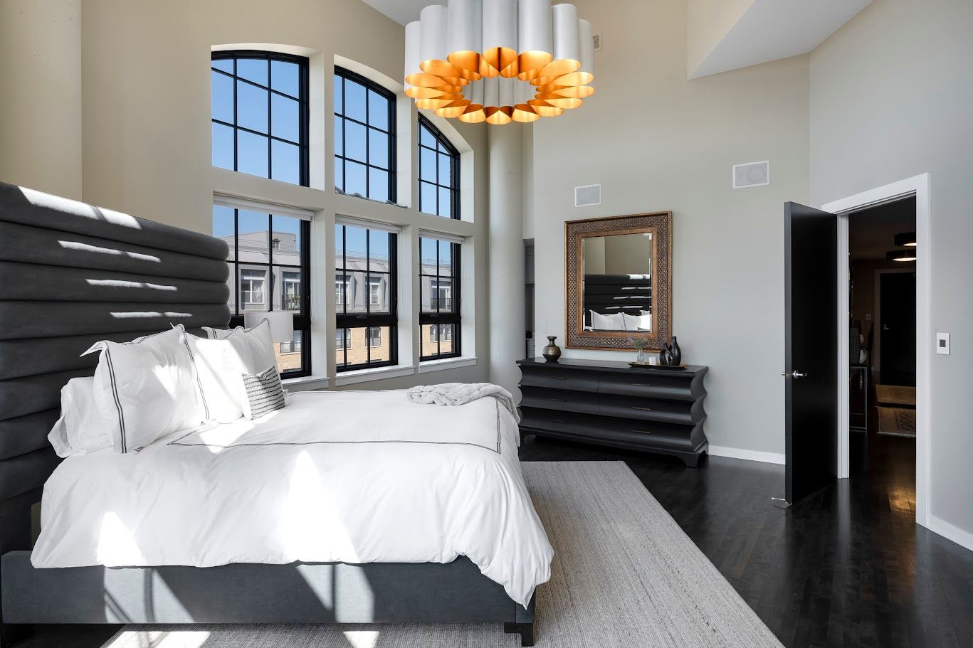 Spacious bedroom with tall windows, high ceiling, dark bed, and dresser. Light-colored rug, modern chandelier.