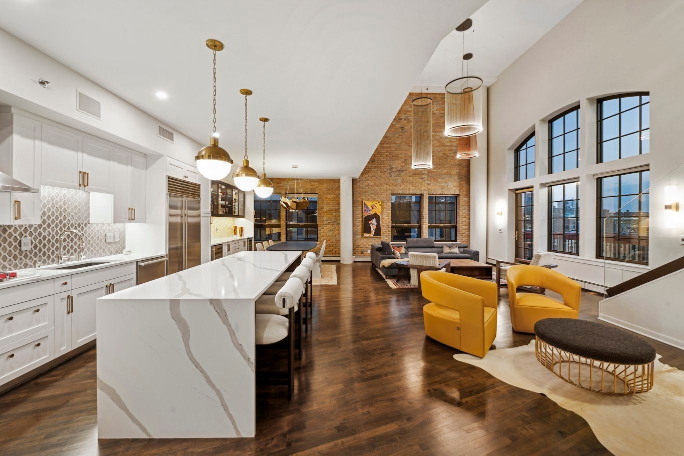 Modern open-concept living space with kitchen island, yellow chairs, exposed brick wall, and tall windows.