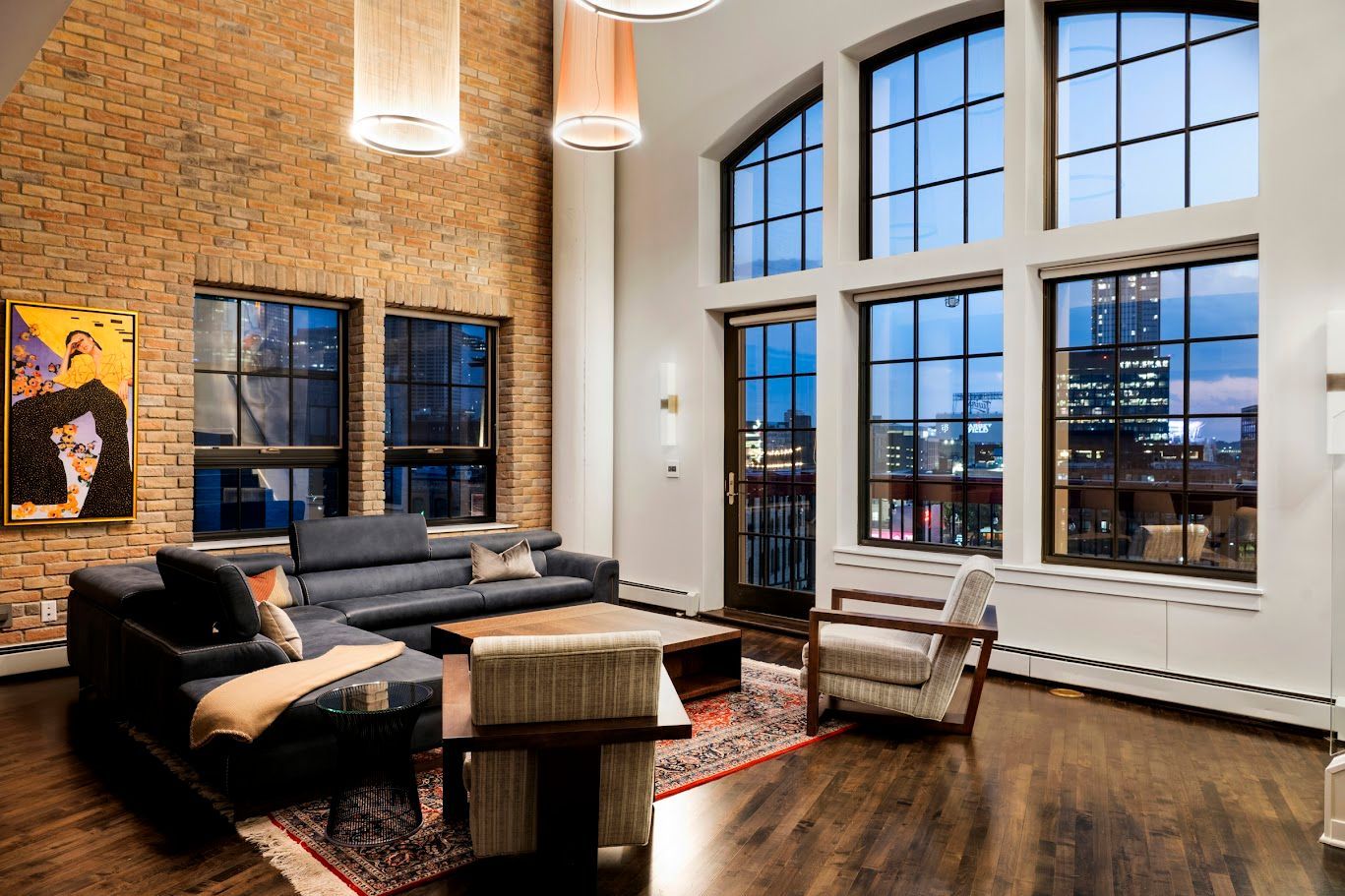 Loft living room with large windows, exposed brick wall, dark wood floors, and blue sofa.