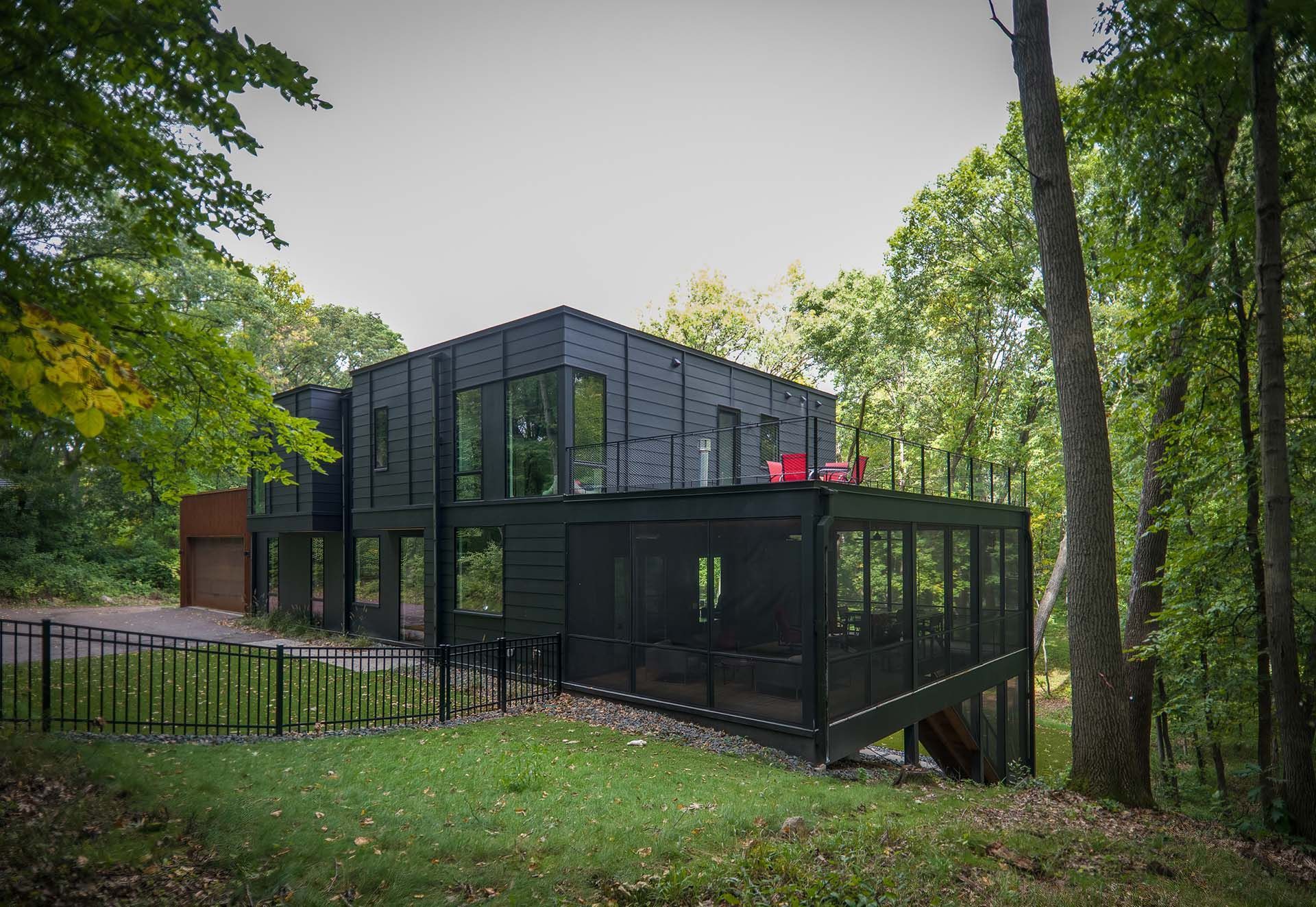 A black house with a large deck is surrounded by trees.