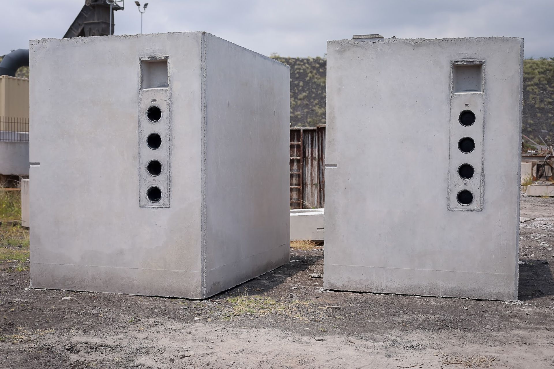 Two small concrete utility structures with vertical circular vents in an outdoor gravel area.