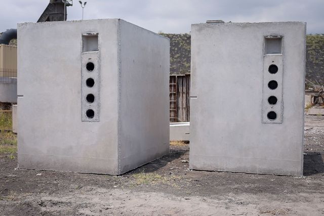 Two white concrete boxes with vertical vents and circular openings, standing outdoors on gravel.