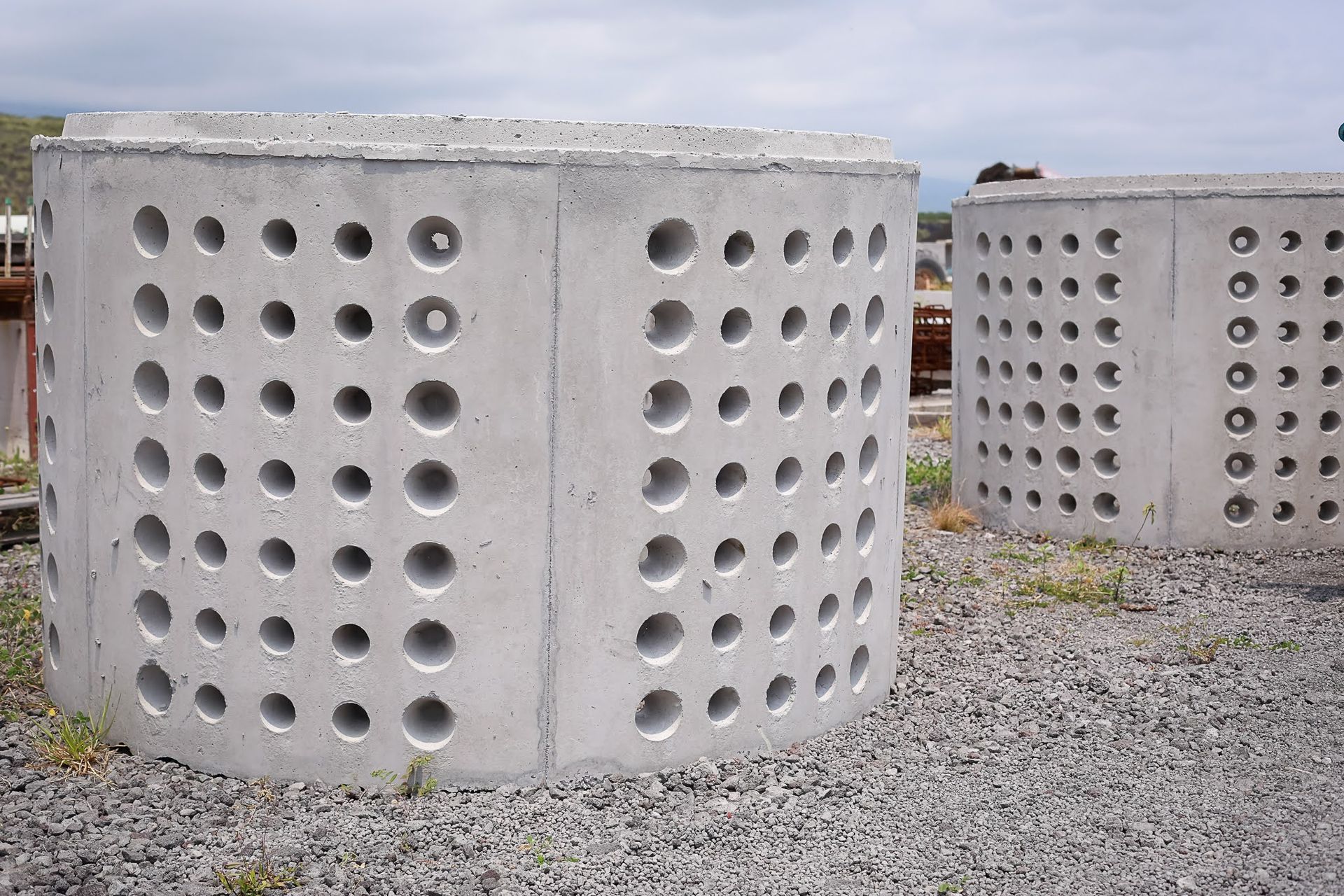 Large concrete cylindrical structures with rows of round holes on a gravel lot under an overcast sky