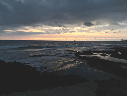 Dark rocky shoreline at dusk with waves under a cloudy sky and a pale sunset on the horizon