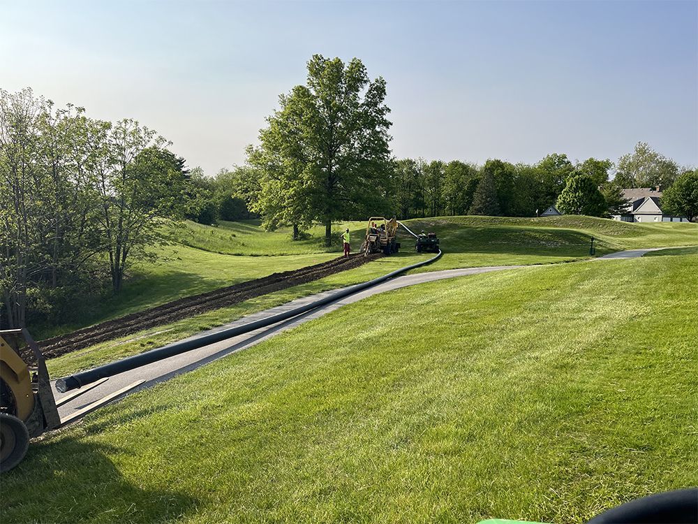 A black pipe being laid across a grassy landscape by a yellow tractor, under a blue sky.