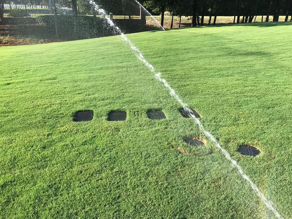 Sprinkler watering green grass on a lawn, with several square access hatches visible.