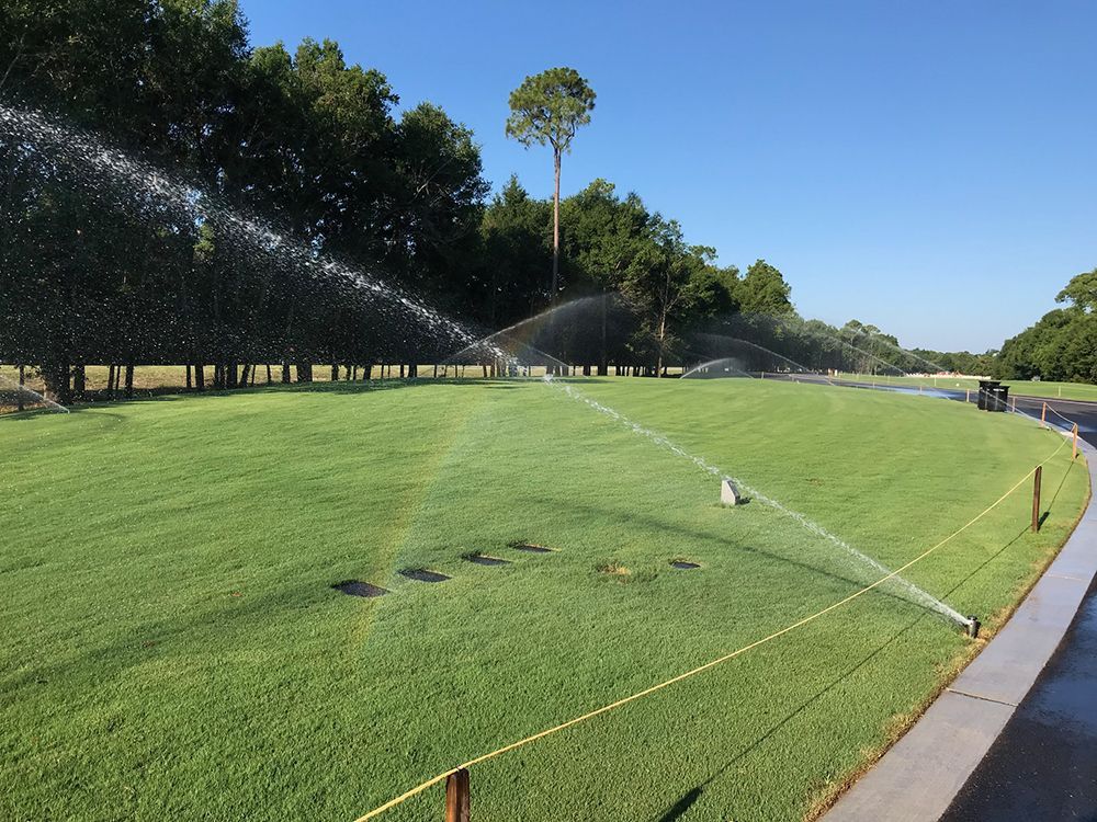 Sprinklers watering green grass on a golf course with trees in the background under a blue sky.