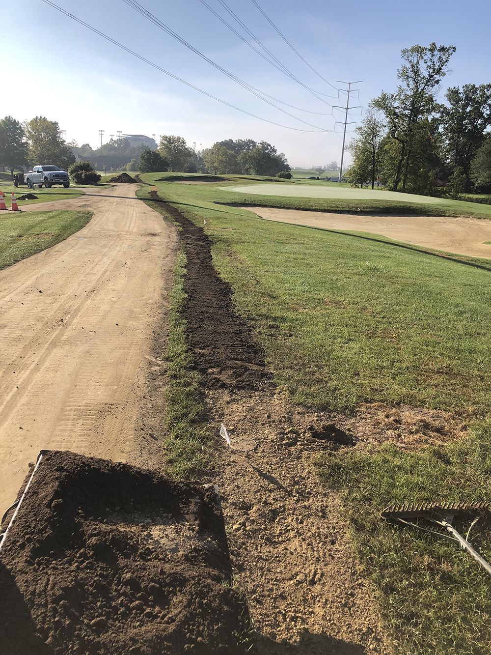 Dirt path and grass under repair at a golf course. Sod on the ground, trees and power lines in the background.