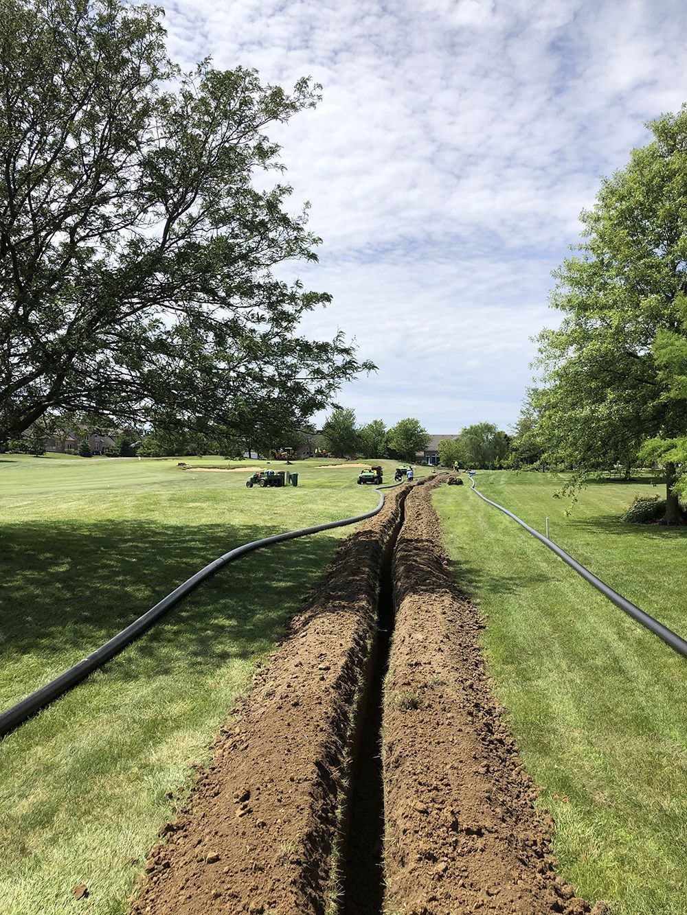 A long trench dug in a green lawn, with black pipes alongside it, under a cloudy sky.