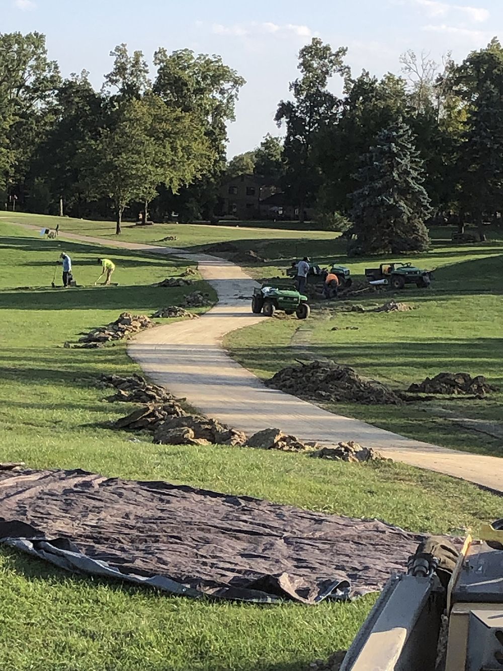 Pathway construction in park; workers, machinery, and dirt piles on grass.