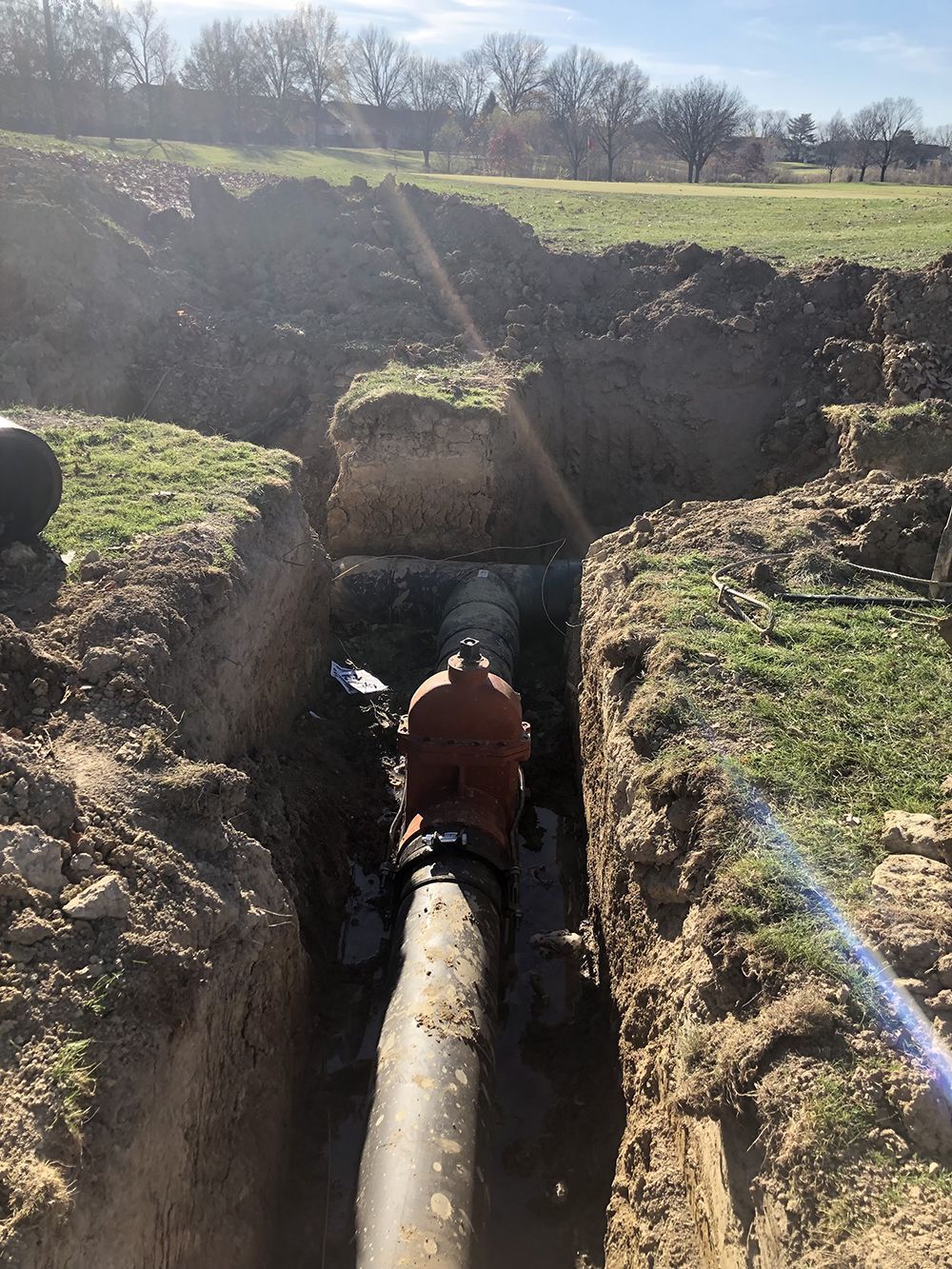 Trench dug around a large pipe with a valve, in a grassy field on a sunny day.