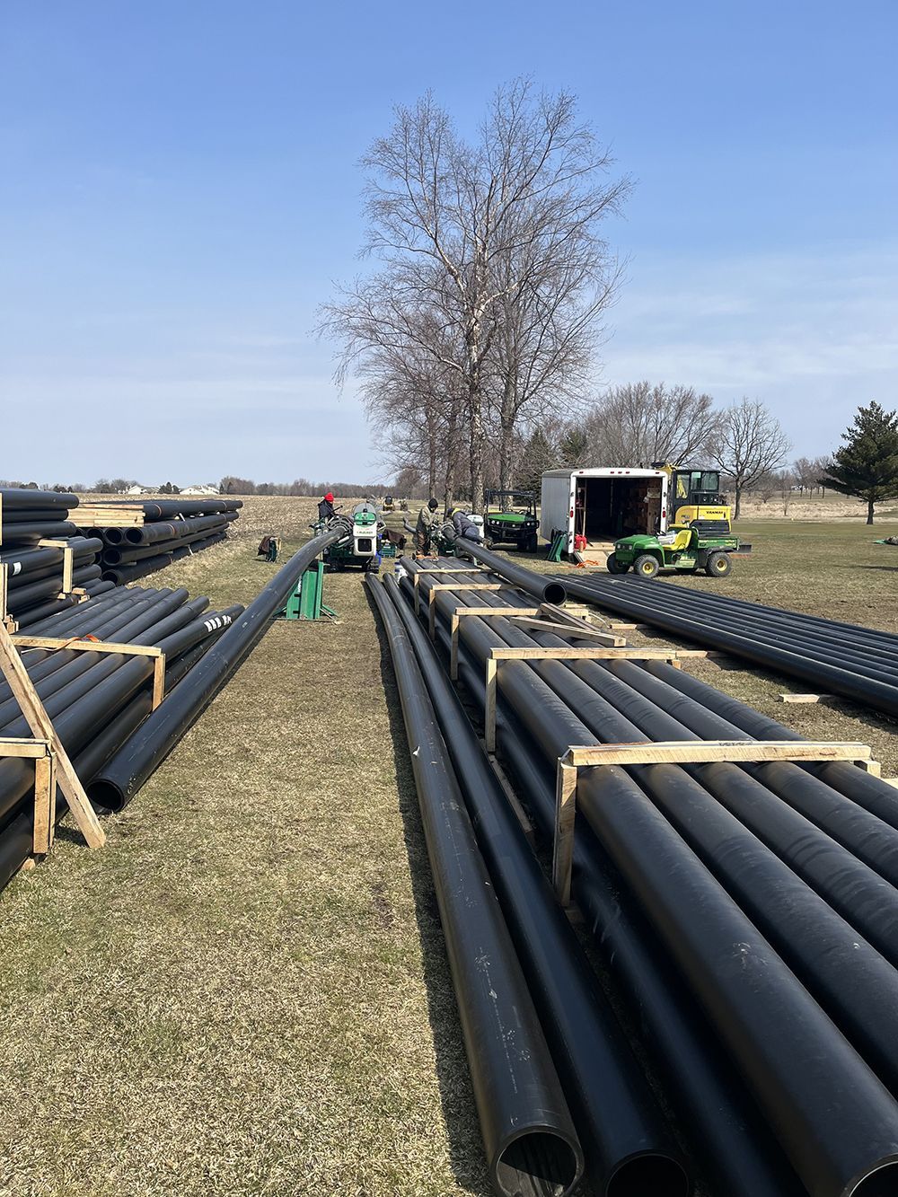 Black pipes laid out on grass field, construction site with machinery and a tree in the background.