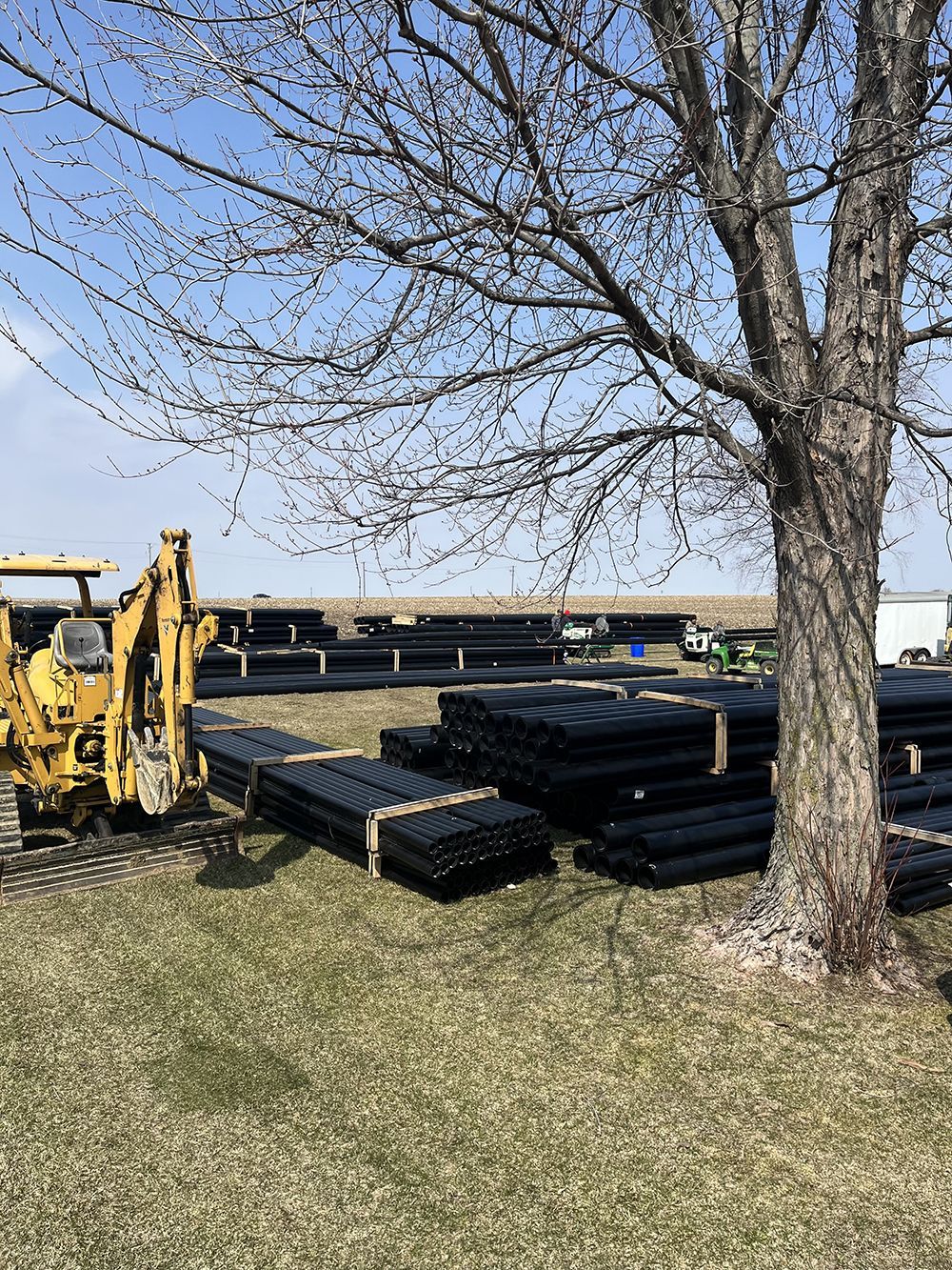 Construction site with black pipes, backhoe, and leafless tree on a grassy field under a cloudy sky.