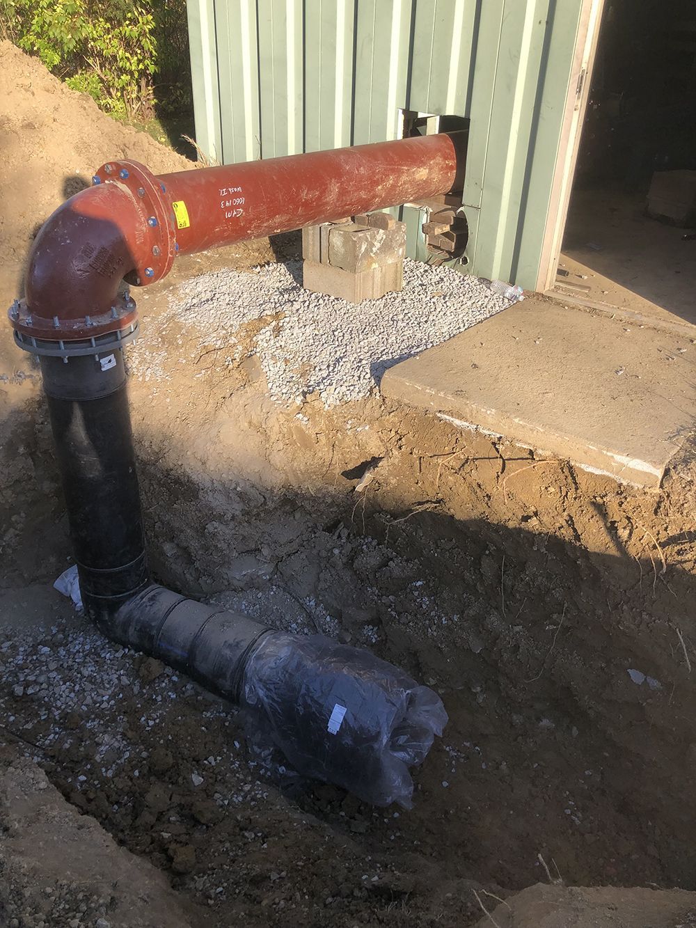 Pipes and fittings in a trench, including a black vertical pipe, a red elbow, and a horizontal red pipe.