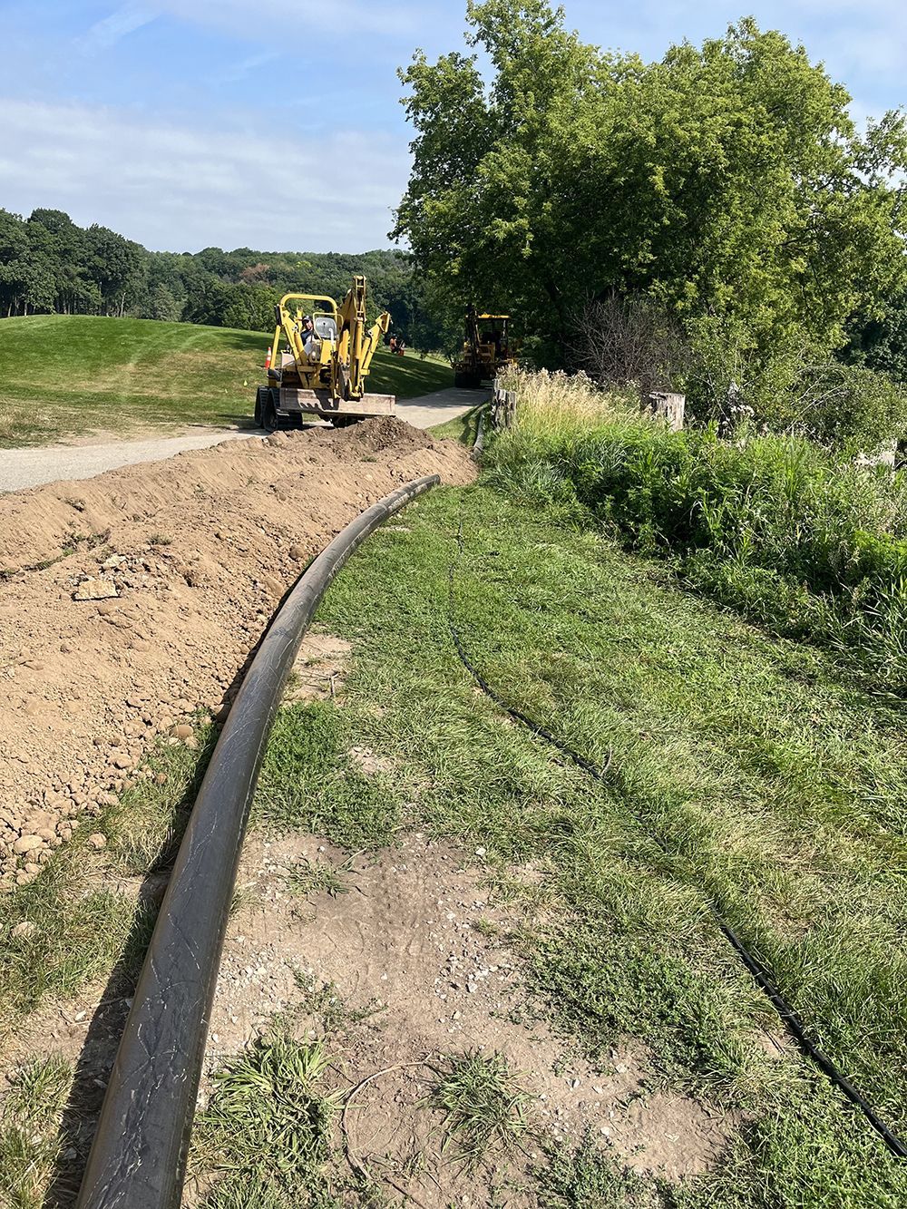 A black pipe lies in a trench alongside grass. A backhoe works in the distance on a gravel path near trees.