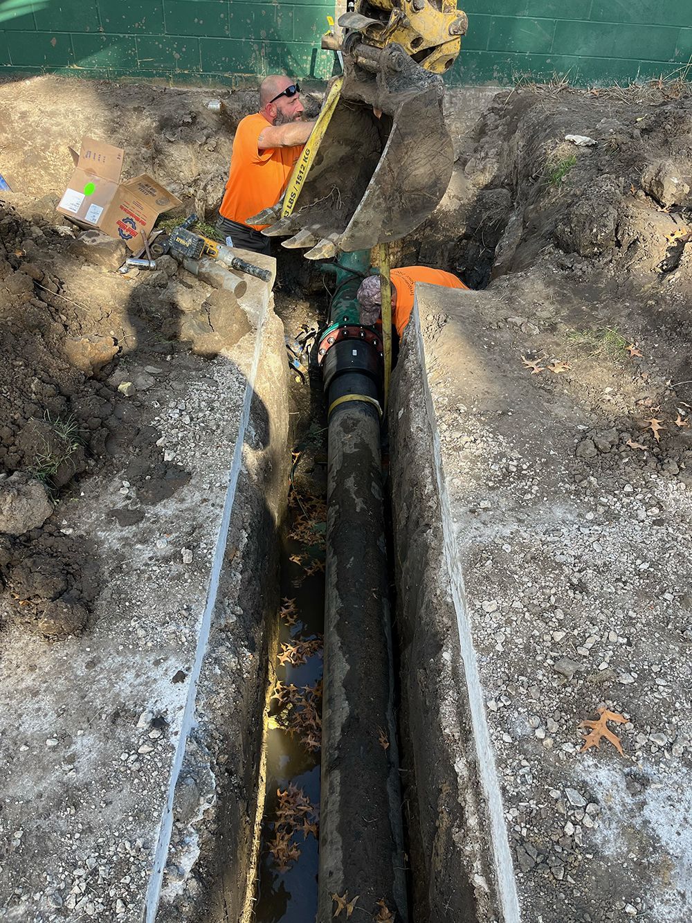 Construction worker with excavator repairing underground pipe in trench.