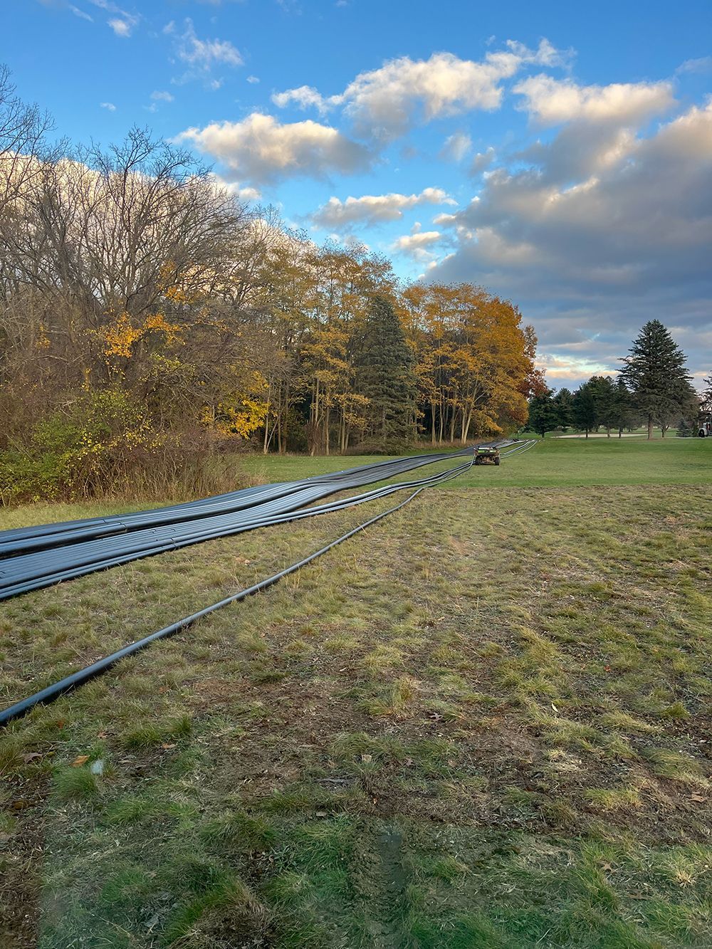 Black pipes laid across grassy field, trees in the background, blue sky with clouds.
