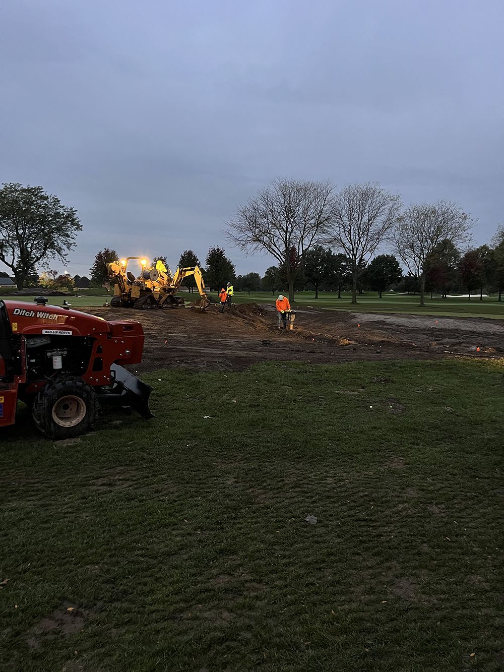Construction site on a golf course at dusk. Ditch Witch, excavator, worker in orange vest.