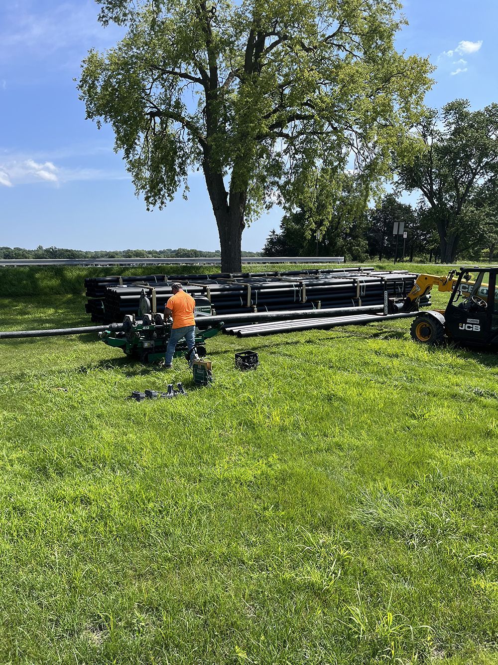 Man assembling stage equipment on green grass near a lake under a tree on a sunny day.