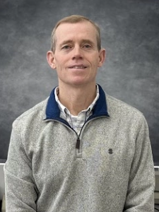 Man in gray sweater smiles, standing in front of a gray backdrop.