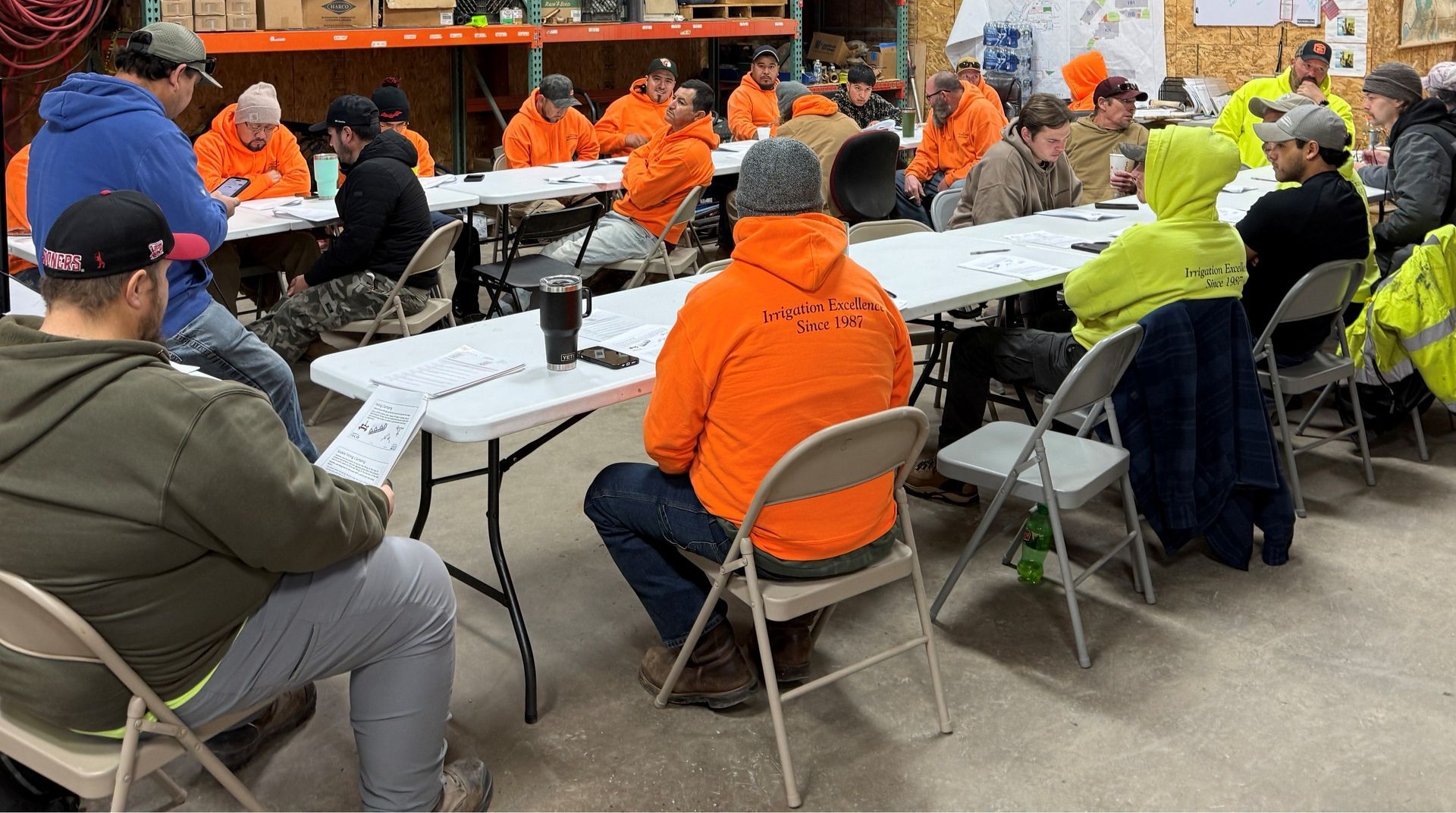 Group of people in a meeting, many wearing orange and yellow safety gear, sitting at long tables in a warehouse setting.