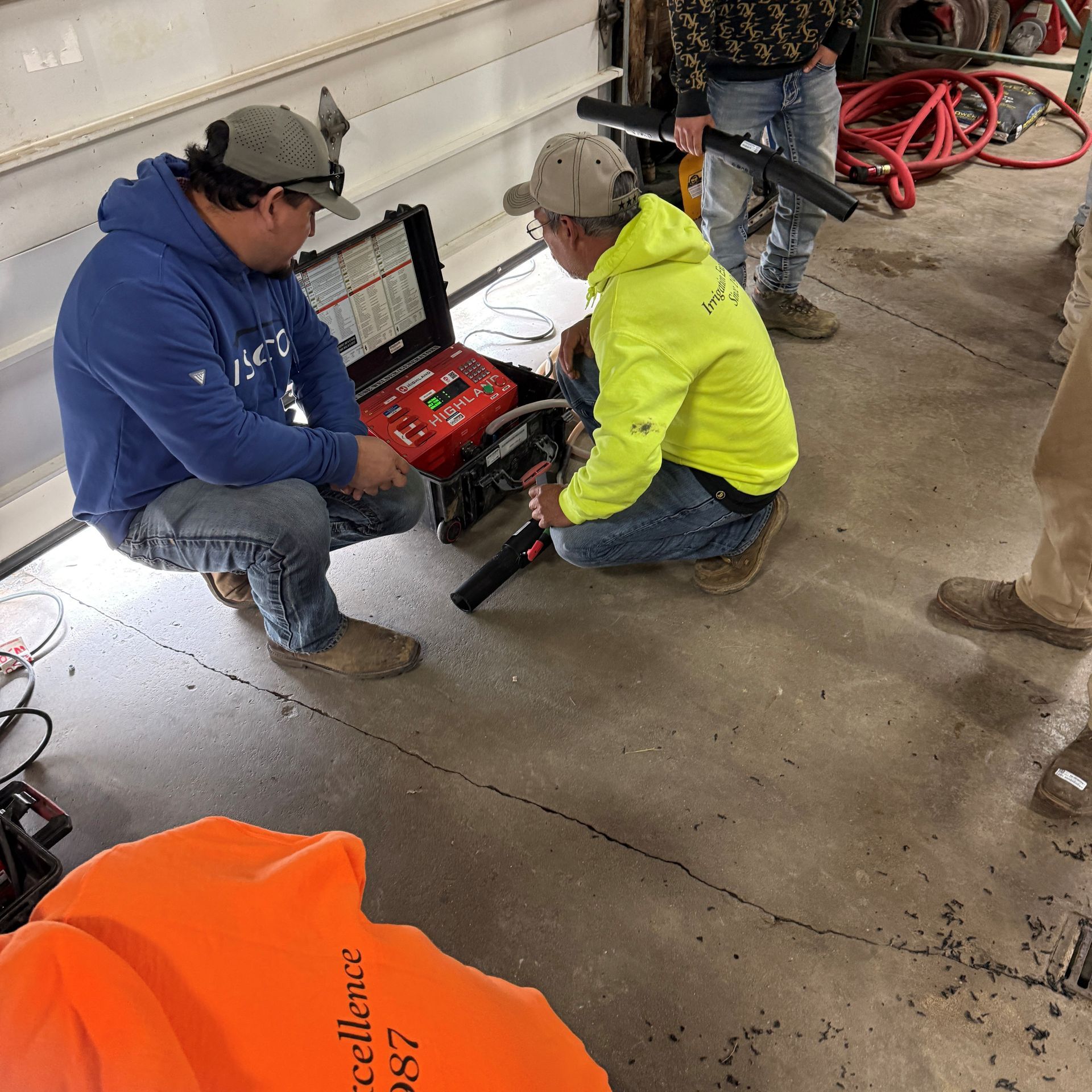 Two people in work clothes inspecting a machine on a garage floor. One holds a black tool.