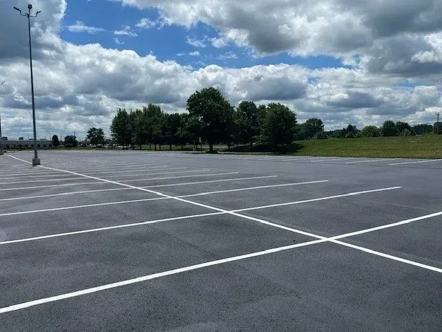 Empty parking lot with white lines under a cloudy sky, trees in the distance.