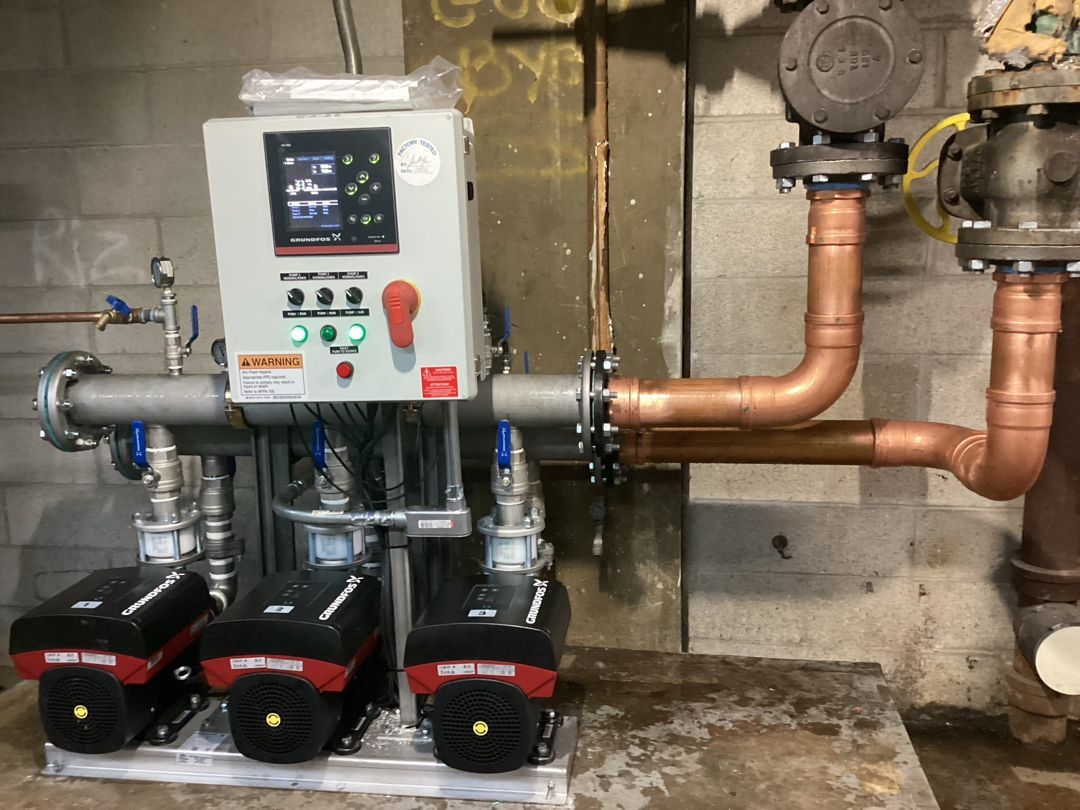Industrial pump control panel with three black pumps and copper piping in a basement-like utility room
