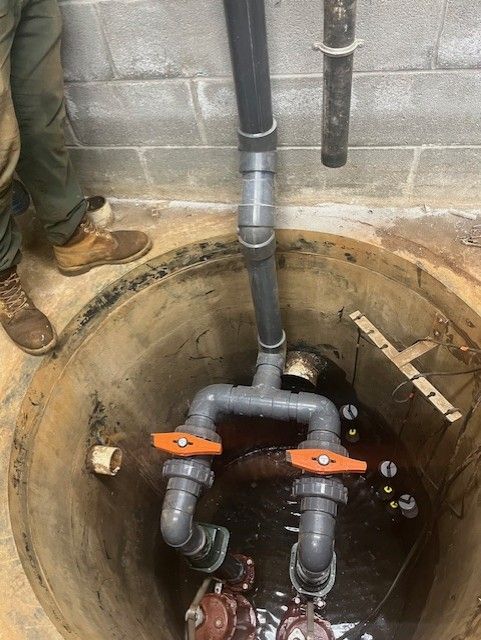 Open utility pit with pipes, orange valves, and standing water in a concrete basement wall opening