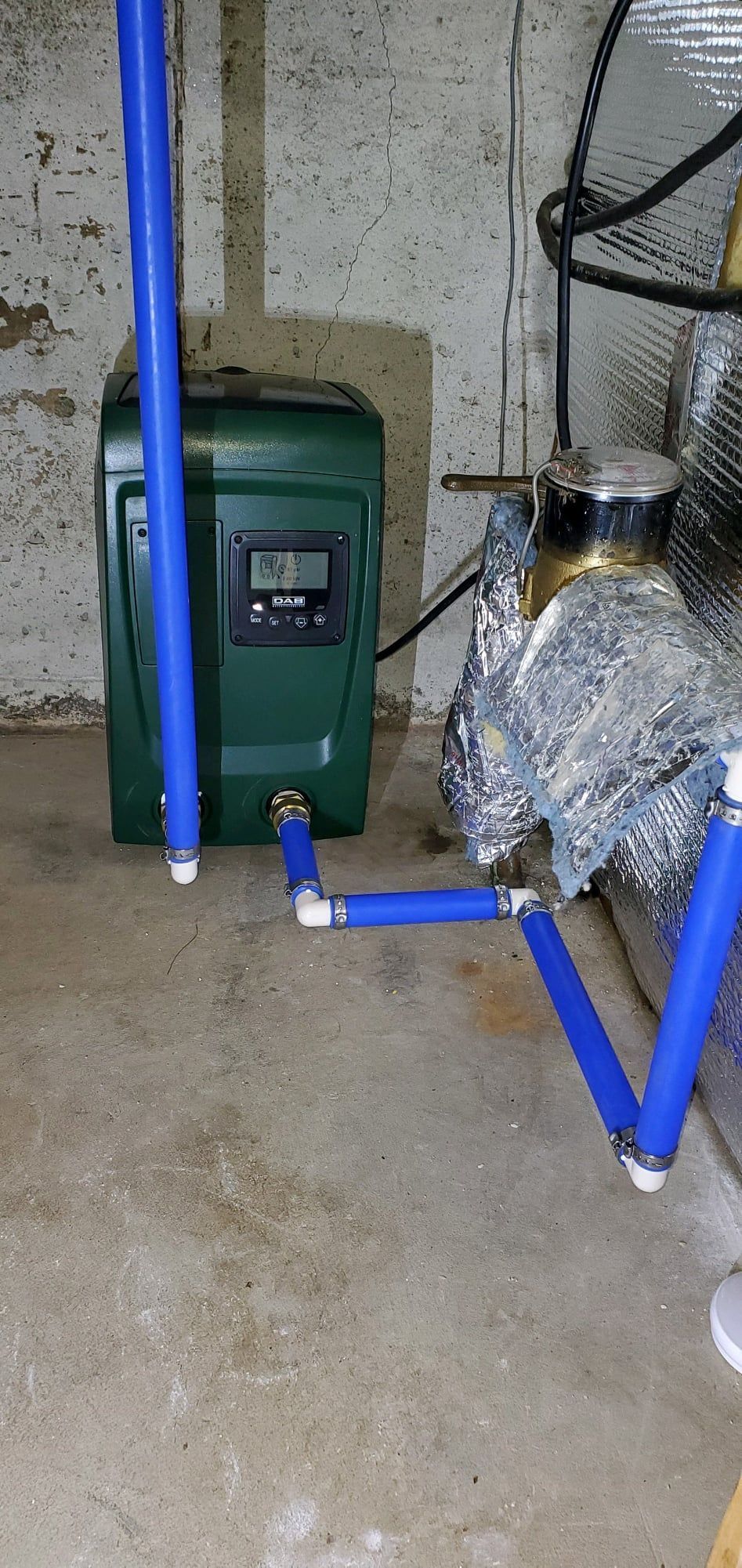 A green water softener unit with a digital display connected to blue PEX piping on a concrete floor next to insulated pipes.