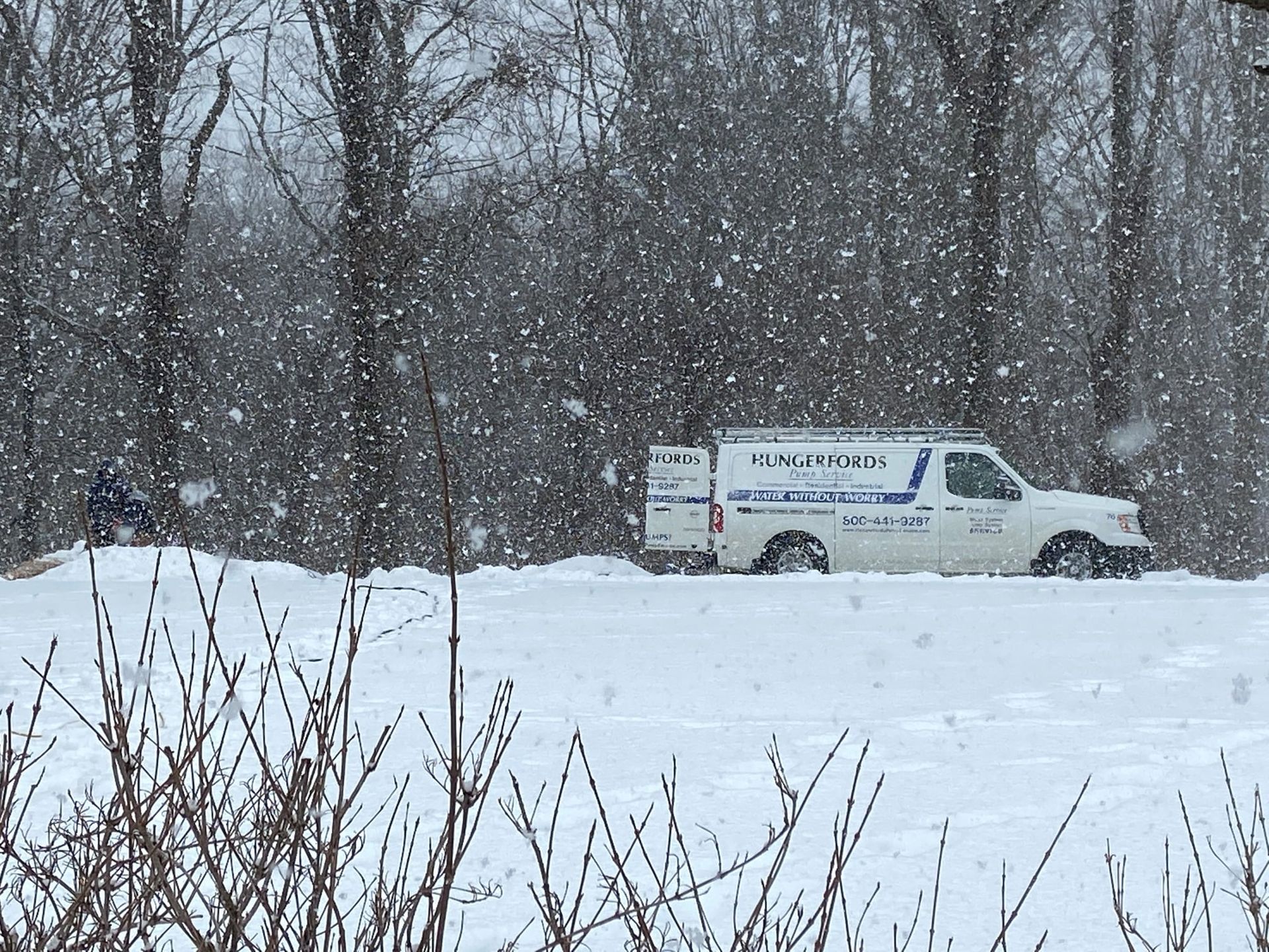 A white service van parked in a snow-covered field during a heavy snowstorm, with trees in the background.