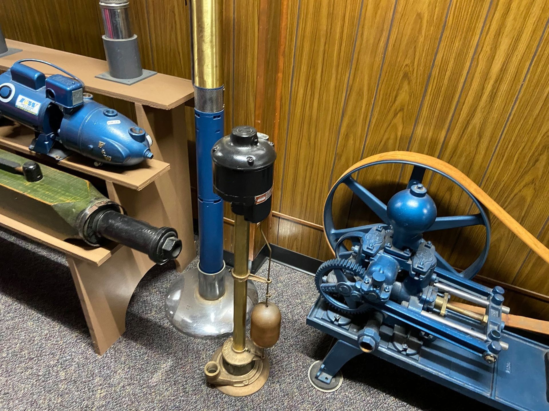 A collection of vintage industrial pumps and machinery displayed on shelves and the floor against a wood-paneled wall.
