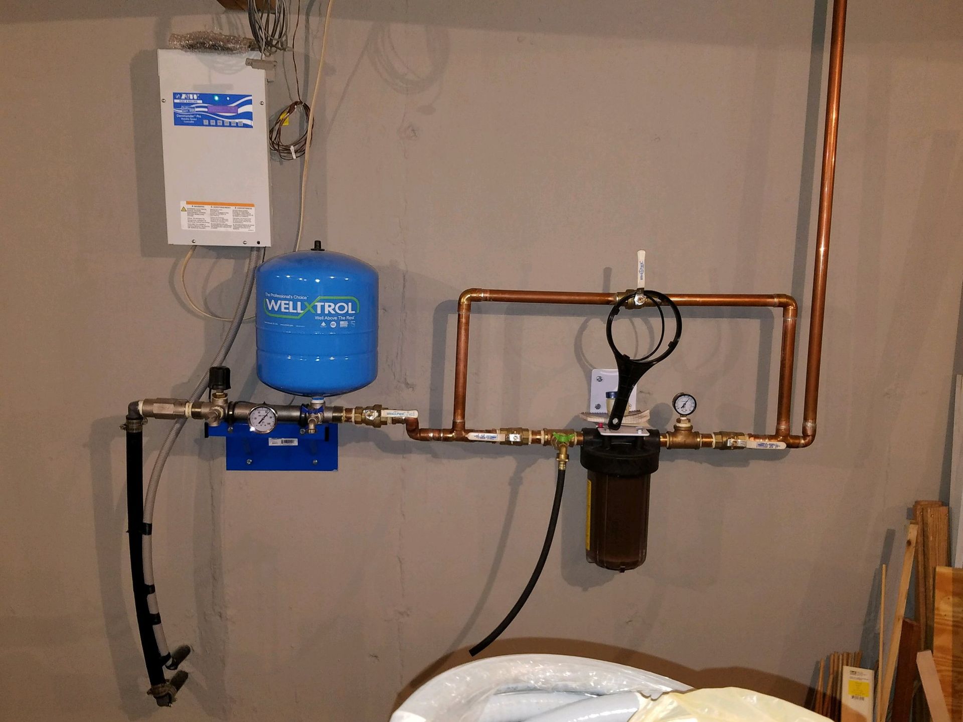 A water filtration system with a blue pressure tank, pipes, and a filter cartridge mounted on a gray wall.