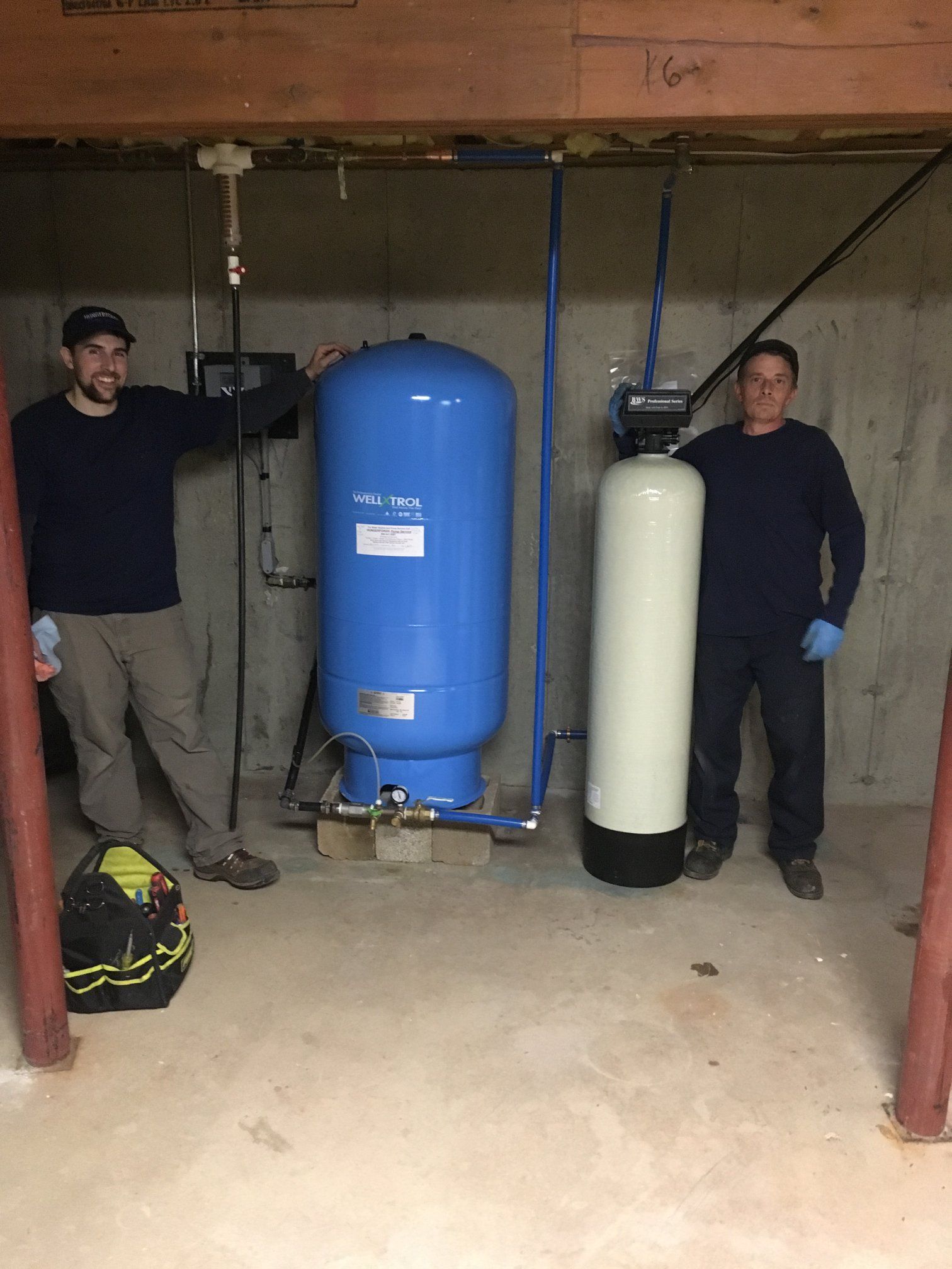 Two people stand in a basement beside a blue water pressure tank and a tall, beige water filtration tank.