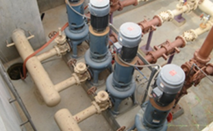 A high-angle view of four vertical industrial pumps connected by a network of pipes in an indoor facility.