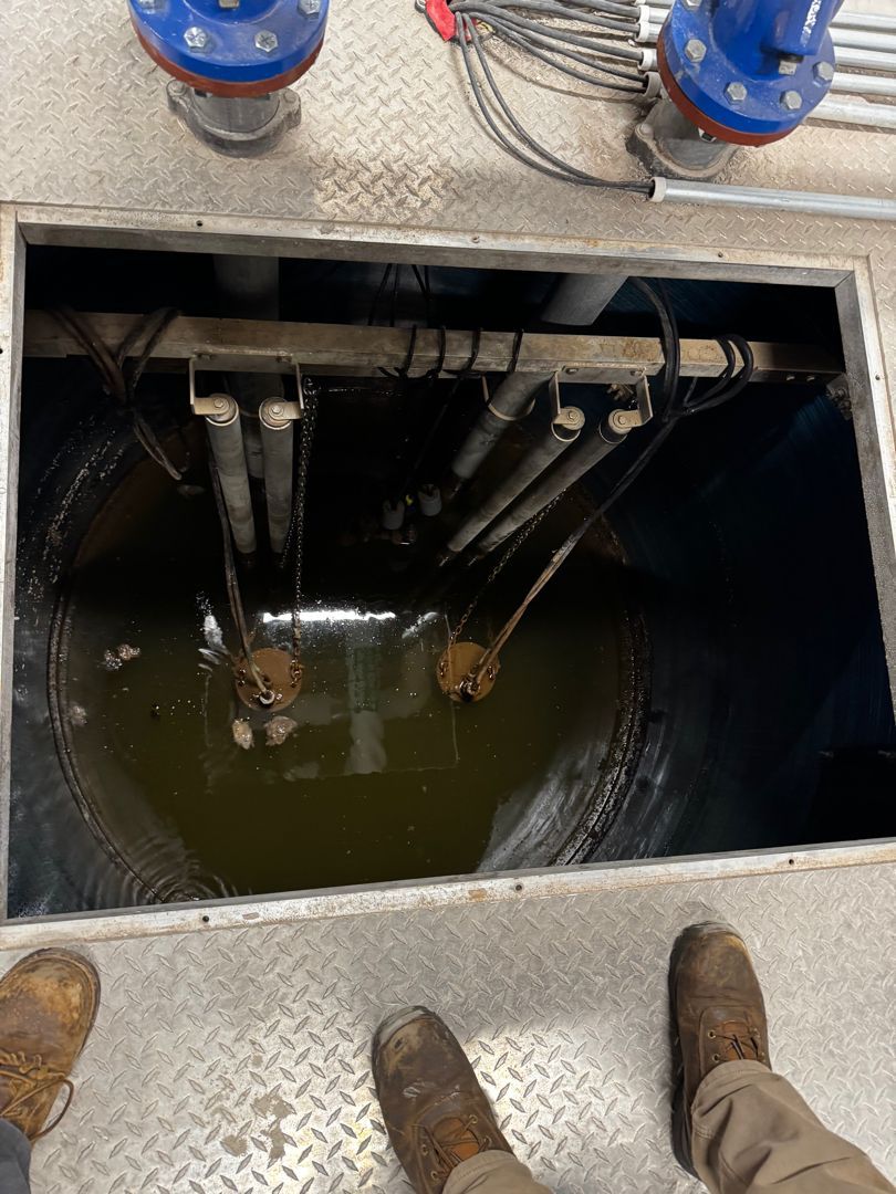 Open hatch in a concrete floor with dark water and pipes inside, boots visible at the edge
