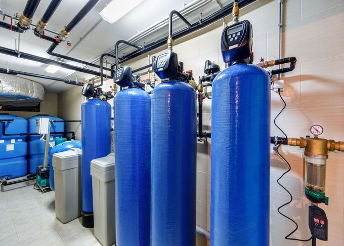 A row of blue industrial water filtration tanks with digital control valves inside a mechanical utility room.