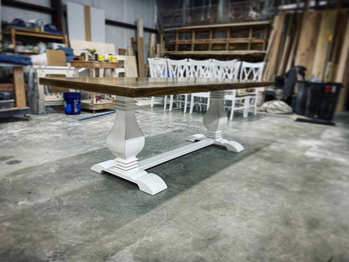 White and brown farmhouse-style dining table. White base, dark wooden tabletop, set in a workshop with chairs in background.