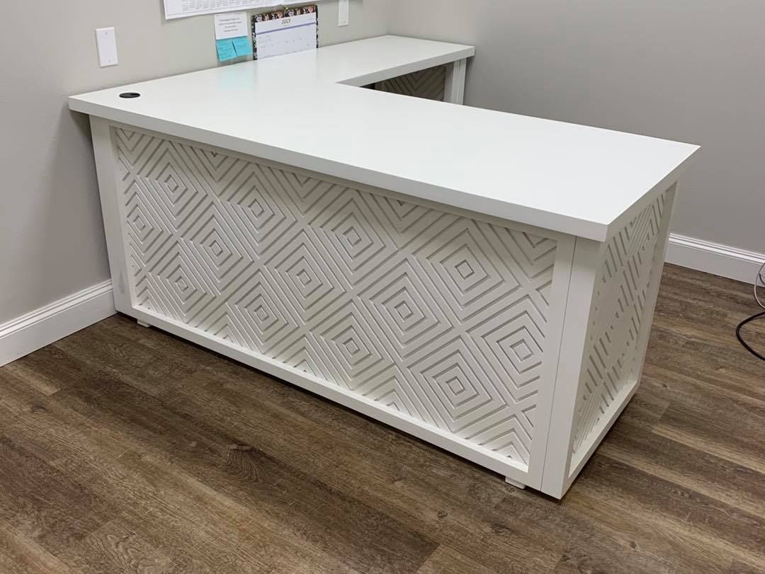 White L-shaped desk with geometric cutouts on the front panel, situated in a corner of a room with wood-look flooring.
