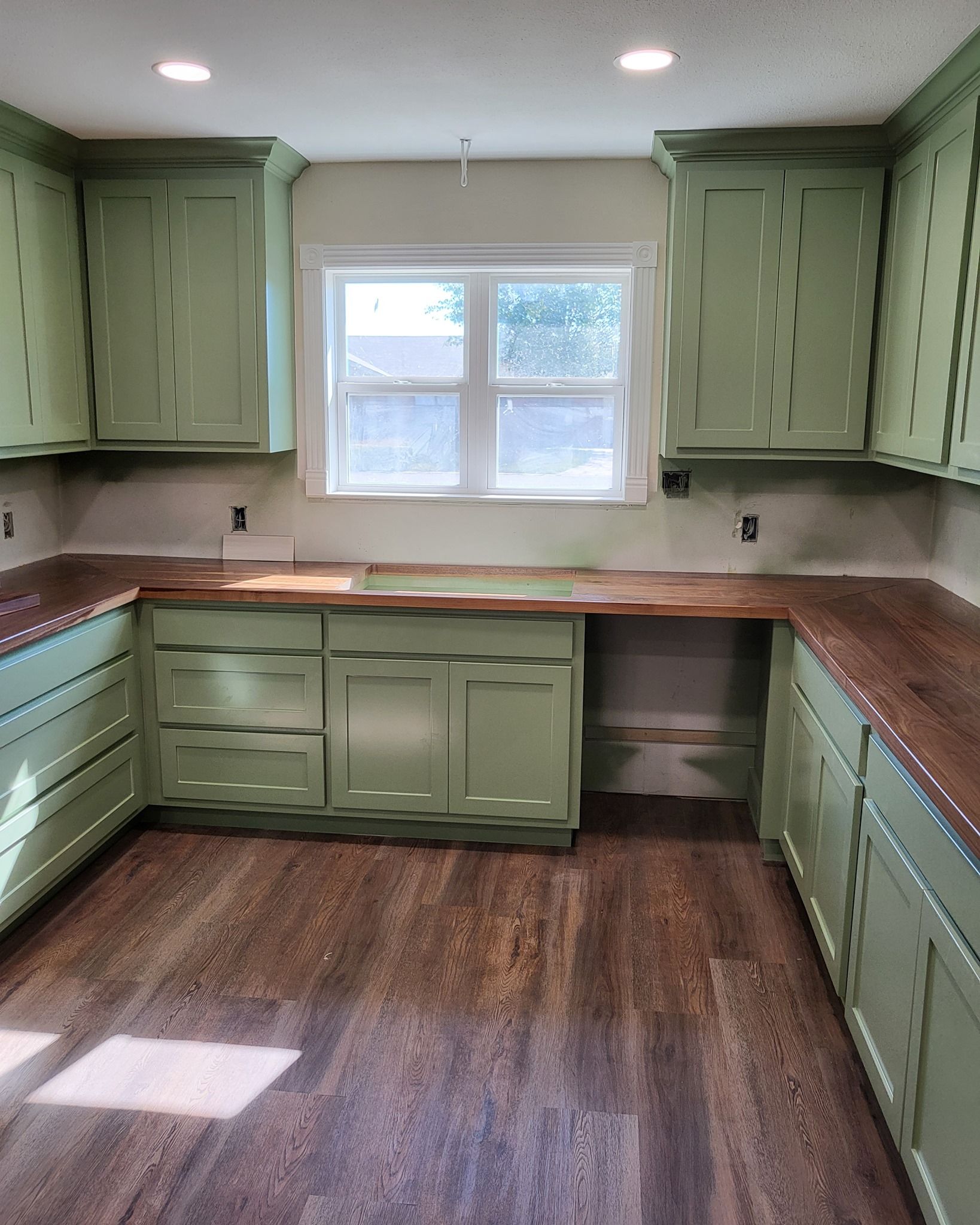 Green kitchen cabinets with wood countertops and flooring. A window is above the open counter space.