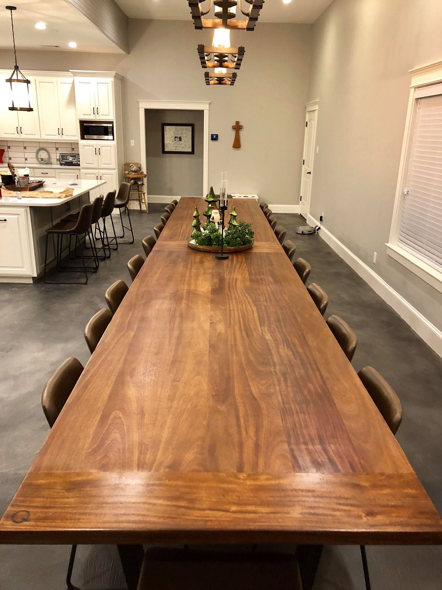 Long wooden dining table set for a gathering in a spacious room. Overhead chandelier, neutral walls, bar seating in the background.