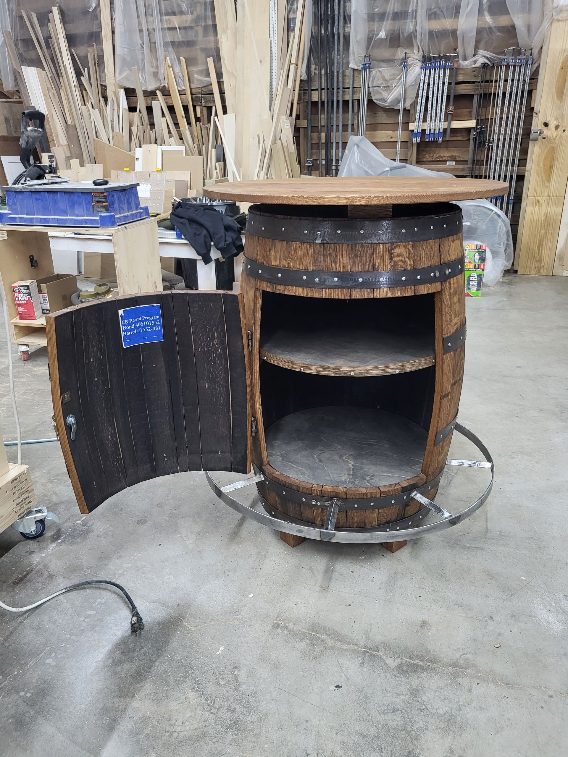 A repurposed wooden barrel bar with shelves and a tabletop, door open, in a workshop setting.