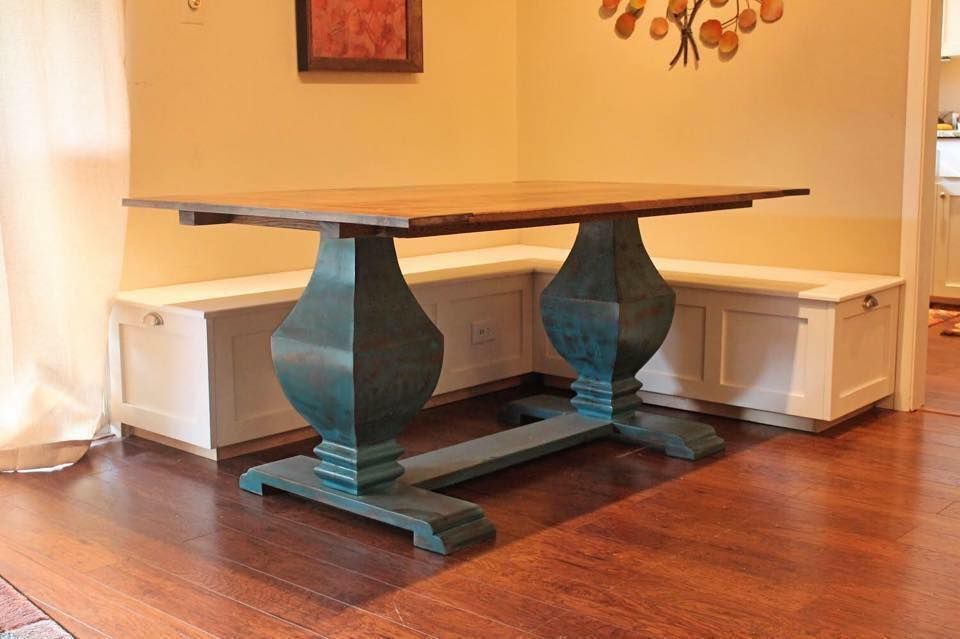 Wooden dining table with teal pedestal base, next to built-in white benches in a room with hardwood floors.