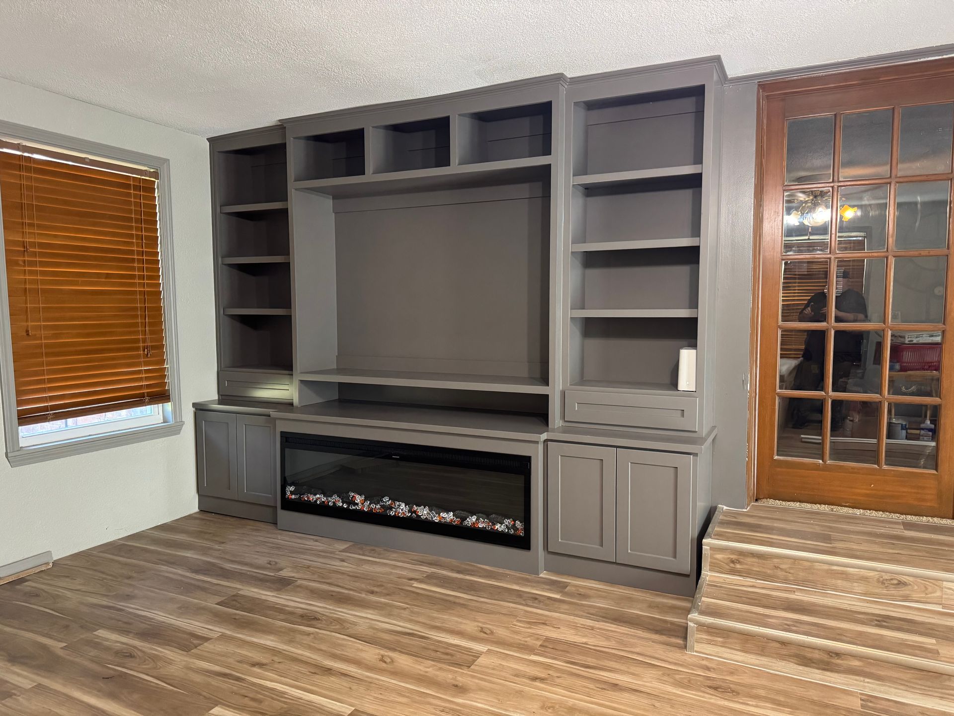 Built-in gray entertainment center with fireplace, shelves, and cabinets. Wooden blinds and glass door with wood trim in the background.